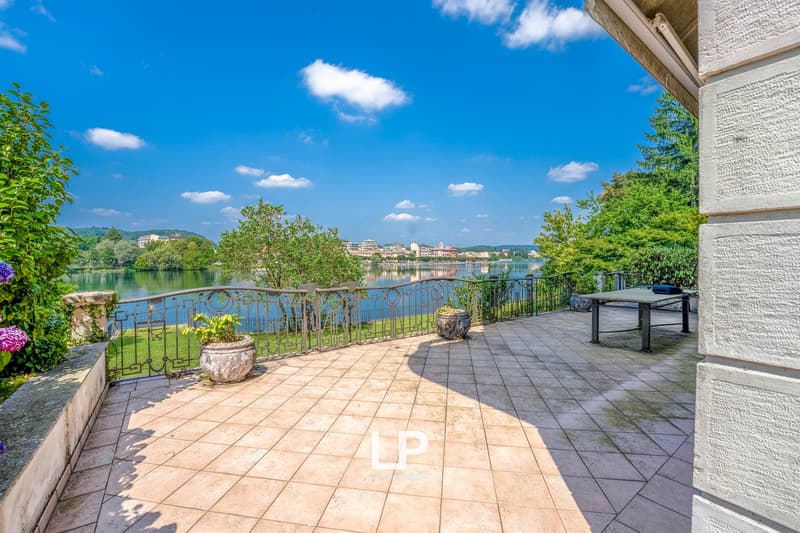 The image shows an outdoor terrace or balcony overlooking a lake or river. The terrace has a tiled floor and decorative railings, with lush greenery and trees surrounding the area. There is a scenic view of the water and buildings in the distance under a