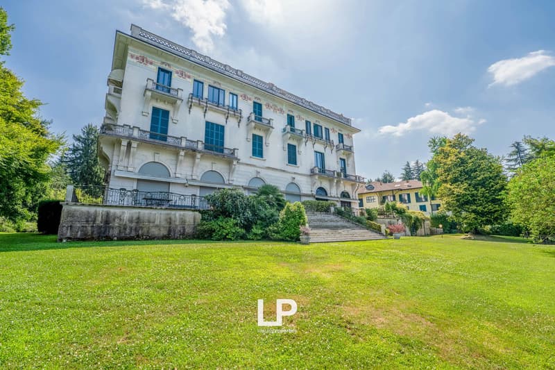 A large, multi-story historic building with a classical architectural style, featuring balconies, arched windows, and a grand staircase leading up to the entrance. The building is surrounded by a well-manicured lawn and lush greenery, creating a picturesq