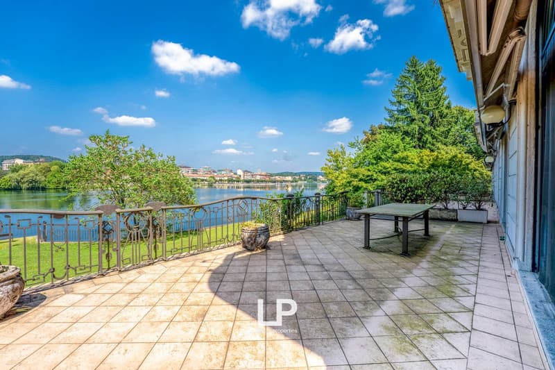 2-story building with a balcony overlooking a lake, surrounded by trees and greenery. The balcony has a tiled floor and metal railing, and there is a table and chairs on the balcony. The view from the balcony includes the lake, buildings, and a blue sky w
