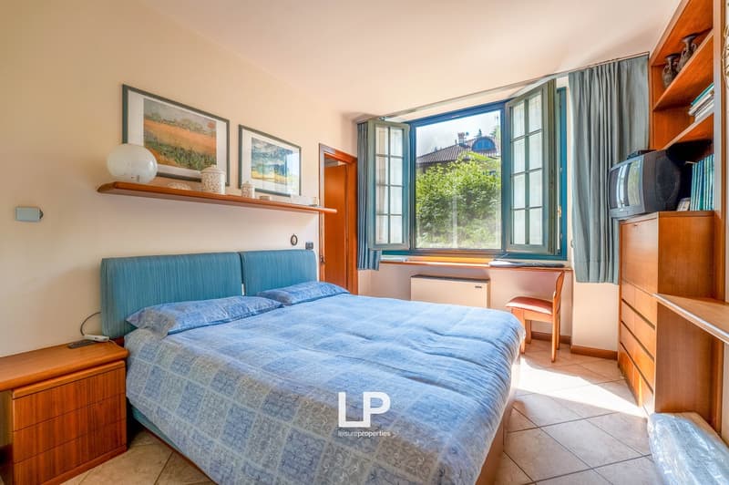 Spacious bedroom with large windows overlooking greenery, blue bedding, wooden furniture including shelves and dresser, and framed artwork on the walls.