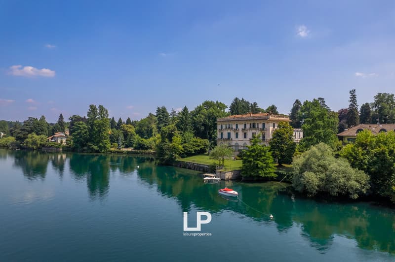 A large, multi-story building with a red roof surrounded by lush greenery and a lake. There is a small boat on the lake in the foreground.