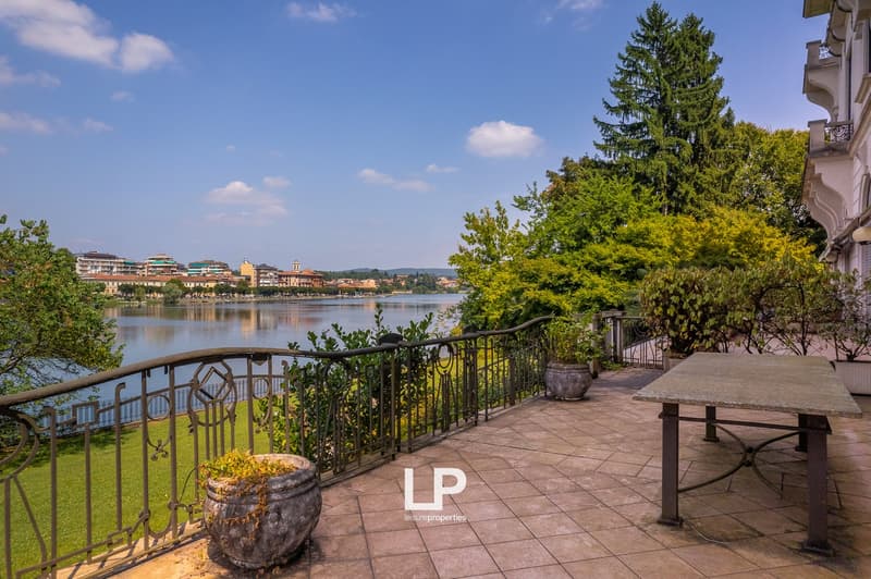 Scenic lakeside view with a paved terrace, ornate railing, and potted plants. The terrace overlooks a lake surrounded by trees and buildings in the distance.