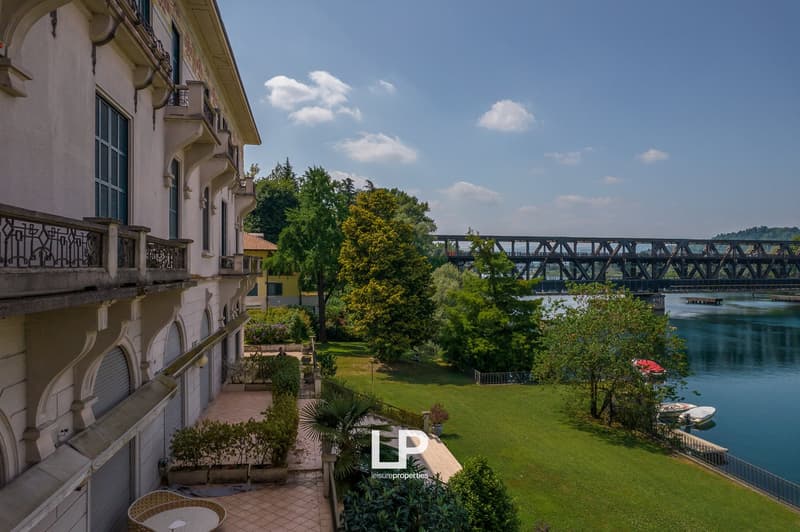 A multi-story building with a balcony overlooking a lush green garden and a river with a bridge in the background. The building has an ornate architectural style with arched windows and a tiled roof.