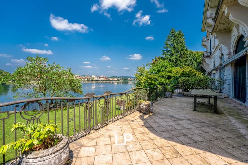Outdoor terrace with ornate railing overlooking a lake, surrounded by lush greenery and a historic building. The terrace has a tiled floor and benches, providing a scenic view of the water and cityscape in the distance.