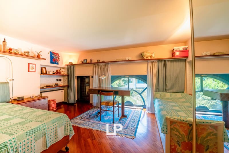 Cozy studio apartment with hardwood floors, large windows providing natural light, a kitchenette area, and a dining table. The room is decorated with various shelves, artwork, and personal items, creating a warm and inviting atmosphere.