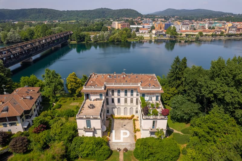 Large multi-story building with red tile roof, surrounded by lush greenery and a lake. The building has balconies and a parking area in front. In the background, there is a city skyline with mountains in the distance.