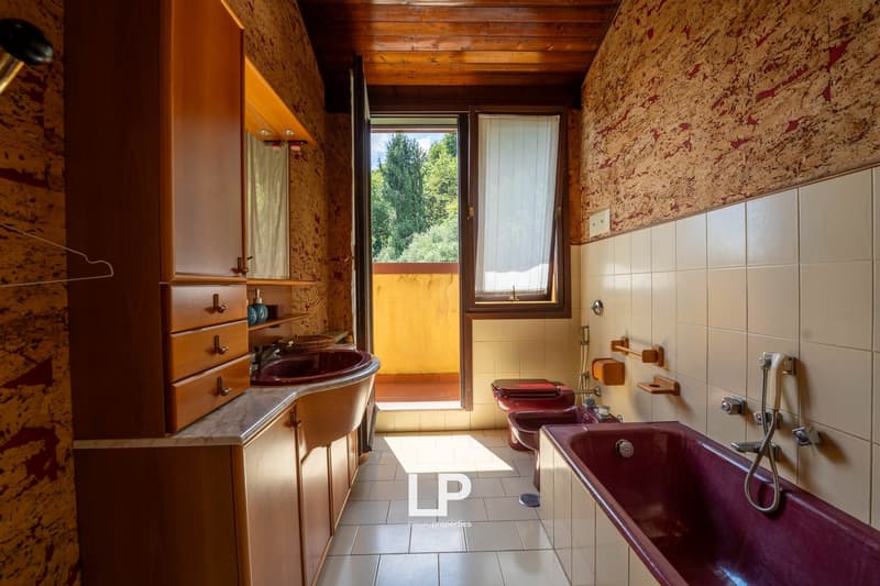 Rustic bathroom with wooden walls and ceiling, large vanity with red sink, tiled floor, and a window overlooking the outdoor scenery.