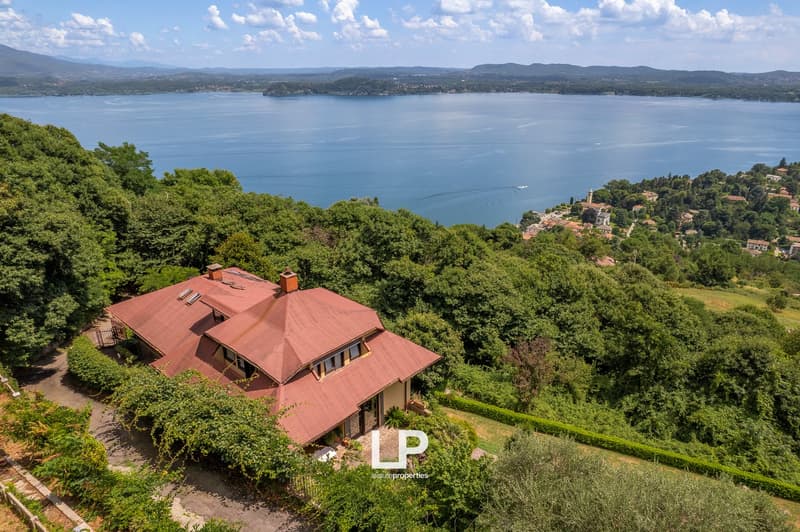 Large two-story house with a red tile roof surrounded by lush greenery, overlooking a scenic lake and mountains in the distance.