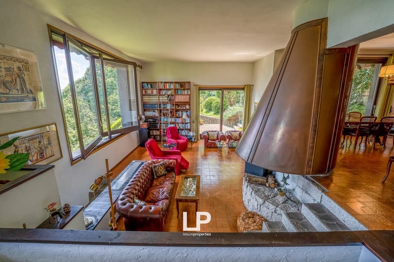 Spacious living room with large windows overlooking the surrounding greenery, built-in bookshelves, red leather armchairs, a tufted leather sofa, and a stone fireplace