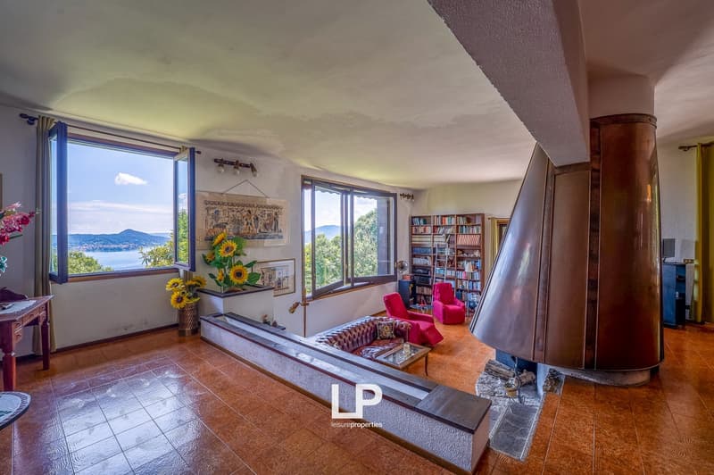 Spacious living room with large windows offering a scenic view of the surrounding landscape, including a lake or body of water. The room features a fireplace, bookshelves, and comfortable seating areas. The decor includes artwork, potted plants, and other