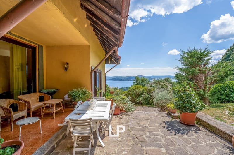 Rustic stone patio with wicker furniture, overlooking a scenic lake and mountains in the distance. The house has a yellow exterior with a tiled roof and wooden beams. The patio is surrounded by lush greenery and potted plants.