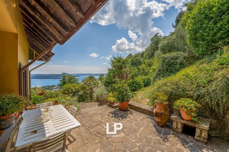 Covered patio with wooden furniture, surrounded by lush greenery and overlooking a scenic lake and mountains in the distance
