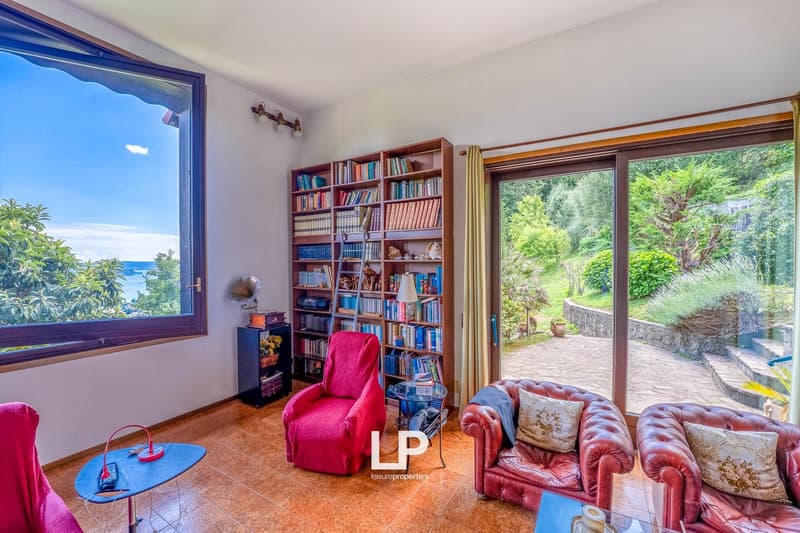 Spacious living room with large windows overlooking a lush, green outdoor area. The room features built-in bookshelves, a red armchair, and a red leather sofa. The floor is made of cork tiles, and the room has a cozy, relaxing atmosphere.