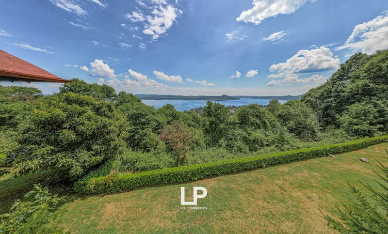 The image shows a scenic view of a lush, green landscape with a large body of water in the distance. The scene features a well-manicured lawn surrounded by dense foliage and trees, with a red-roofed building visible in the background. The sky is filled wi