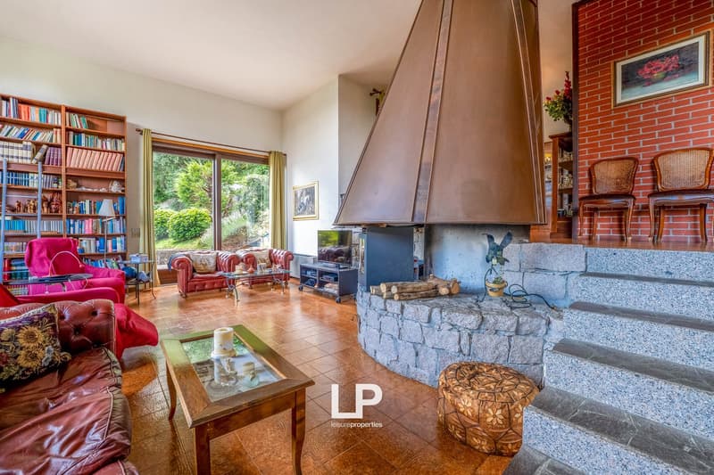 Spacious living room with large windows overlooking the garden, brick fireplace, built-in bookshelves, red leather sofas, and a wooden coffee table.