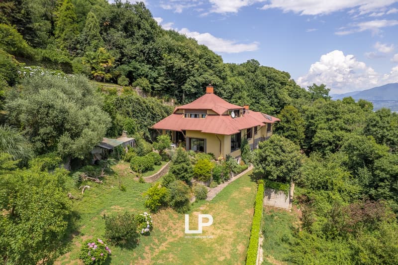 Two-story house with a red tile roof, surrounded by lush greenery and trees, situated on a hillside with a winding path leading up to the property