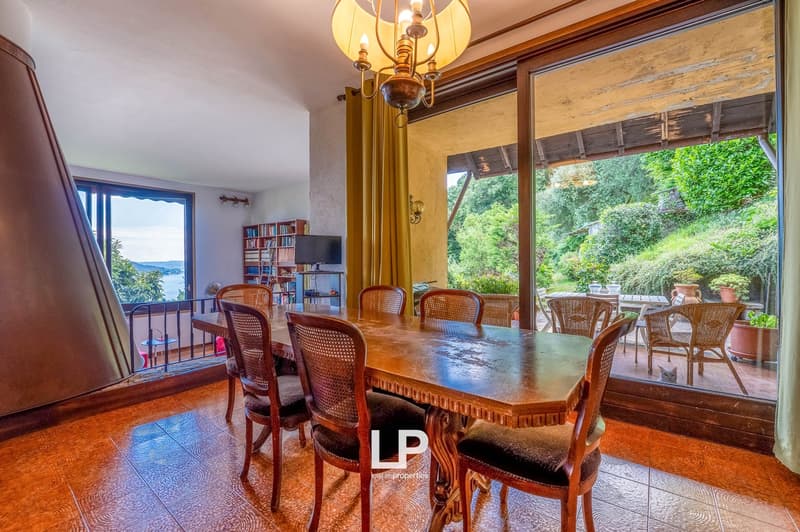 Spacious dining room with large windows overlooking a lush, green outdoor area. The room features a wooden dining table with cane-back chairs, a chandelier, and built-in bookshelves on the side. The overall atmosphere is warm and inviting.