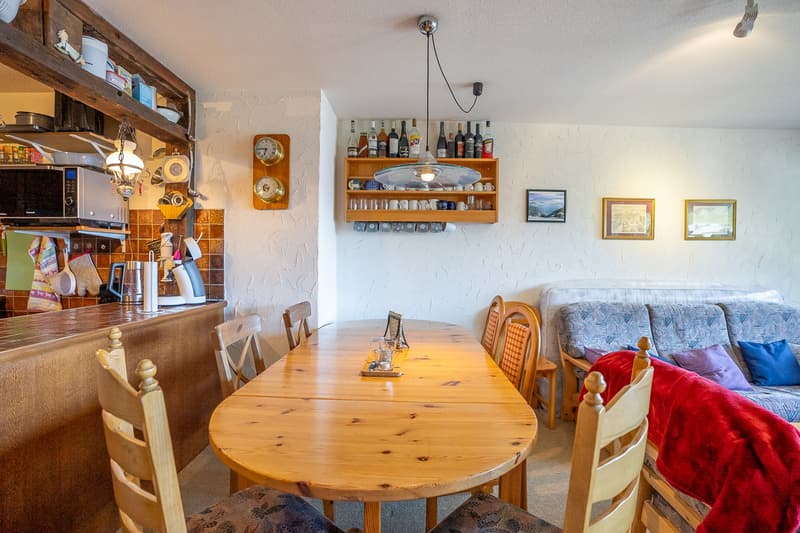The image shows a cozy kitchen and dining area with a wooden dining table, chairs, and a sofa. The kitchen features a tiled backsplash, shelves with dishes and glassware, and various kitchen appliances including a microwave and what appears to be a dishwa