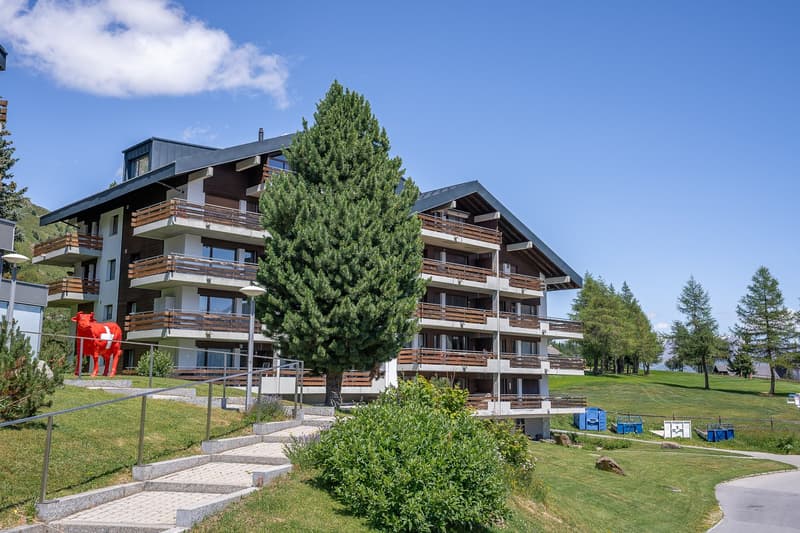 Multi-story apartment building with wooden accents, balconies, and large windows. The building is surrounded by a grassy area with trees and a red sculpture in the foreground.
