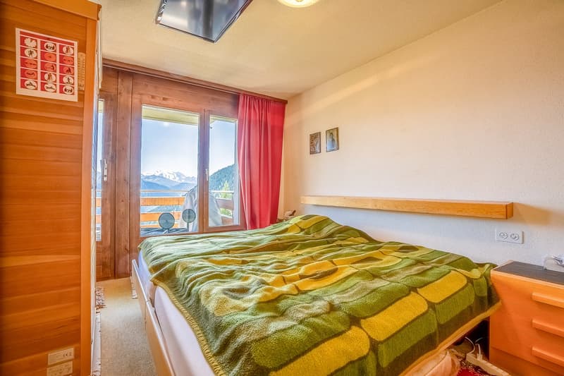 Spacious bedroom with wooden walls, large windows providing a scenic mountain view, a cozy bed with a colorful patterned blanket, and a TV mounted on the wall.