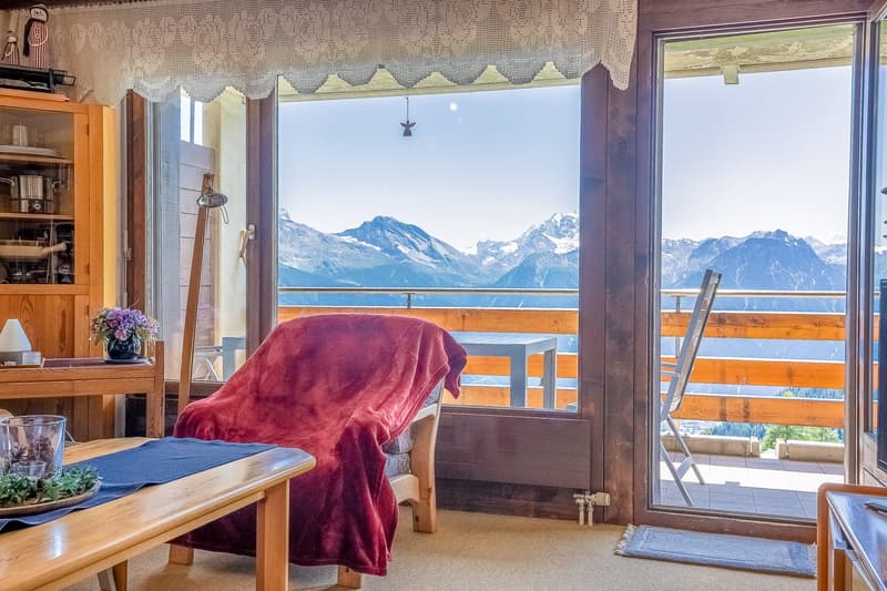 Cozy living room with large windows offering a stunning view of snow-capped mountains, wooden furniture, a red blanket on a chair, and a small table with a potted plant.
