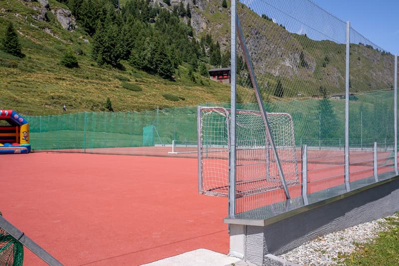 Outdoor sports court with red surface, soccer goal, fencing, and a cable car or gondola in the background on the mountainous landscape