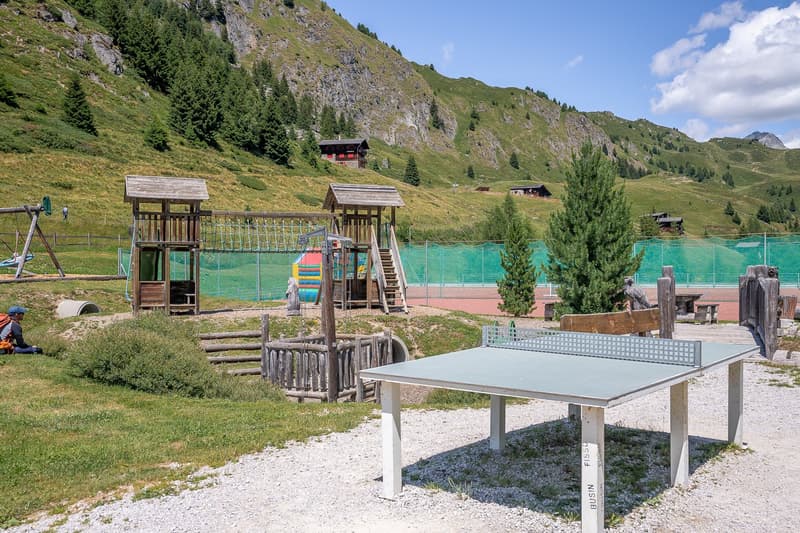 The image shows an outdoor recreational area in a mountainous landscape. There is a playground with wooden structures, a ping-pong table, and a fenced-off area with what appears to be a swimming pool or pond. The area is surrounded by mountains, pine tree