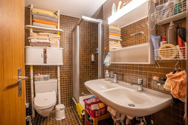 Tiled bathroom with a toilet, sink, and shelves with towels and other bathroom items. The room appears to be well-organized and functional.