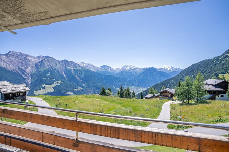 The image shows a scenic mountain landscape with a wooden balcony or deck in the foreground. The view includes snow-capped peaks, lush green forests, and a small village or town nestled in the valley below. The sky is clear and blue, creating a picturesqu