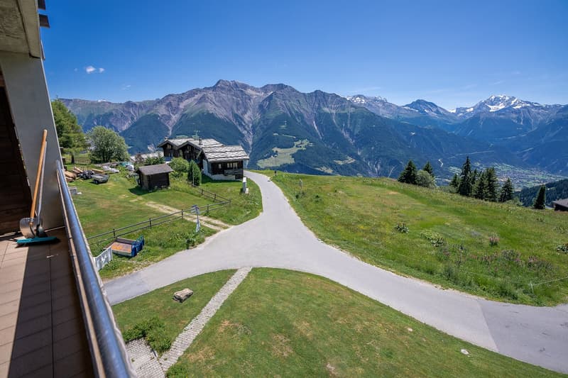 The image shows a scenic mountain landscape with a winding path leading through a grassy area. There are several traditional alpine-style chalets or cabins visible, surrounded by lush greenery and set against a backdrop of towering snow-capped peaks. The
