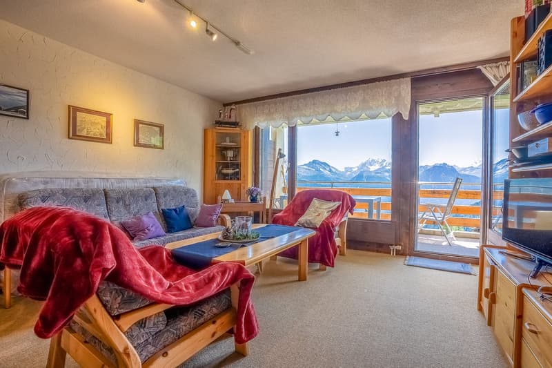 Cozy living room with large windows offering a scenic mountain view, plush gray sofa, red throw blankets, wooden shelving unit, and framed artwork on the walls.