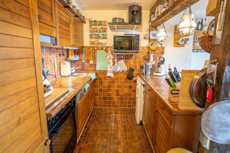 Rustic kitchen with wooden cabinets, tiled floor, various kitchen appliances and utensils, shelves with spices and cookware, and a hanging lamp fixture.