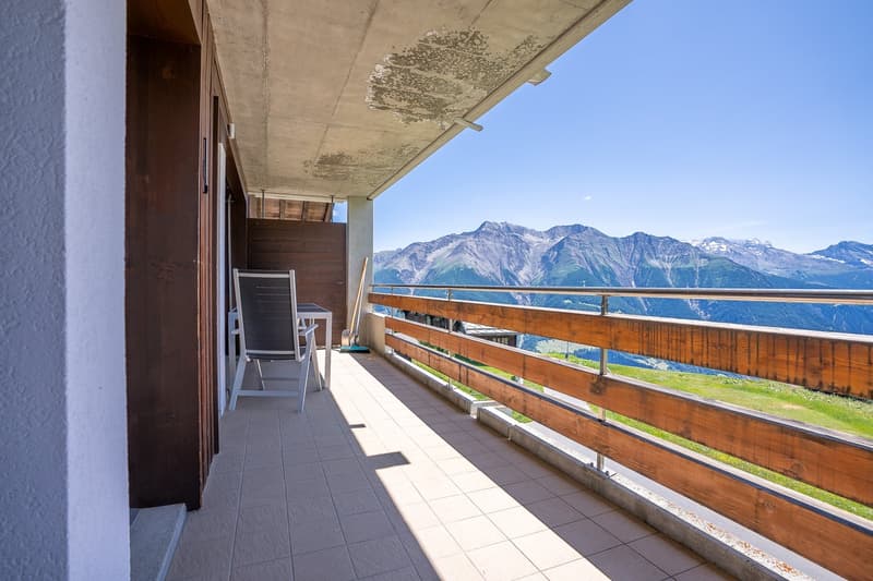 Spacious balcony with a stunning view of the mountains and lake. The balcony has a concrete ceiling, wooden railings, and tiled flooring. There is a chair on the balcony, providing a comfortable spot to enjoy the scenic view.