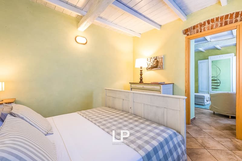 Spacious bedroom with wooden beamed ceiling, yellow walls, wooden furniture, and a checkered bedspread. There is a balcony or terrace visible through the doorway.