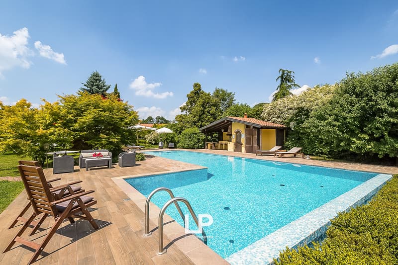 Outdoor swimming pool surrounded by wooden deck, with a small house or cabana nearby. The property is set in a lush, green garden with various trees and vegetation.