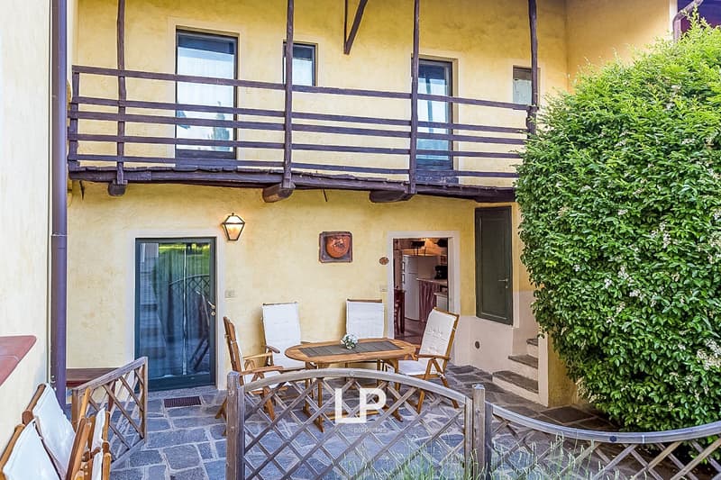 Two-story house with yellow walls, wooden beams, and a balcony with a wooden railing. The balcony has a table and chairs, and there is a small garden area in front of the house.