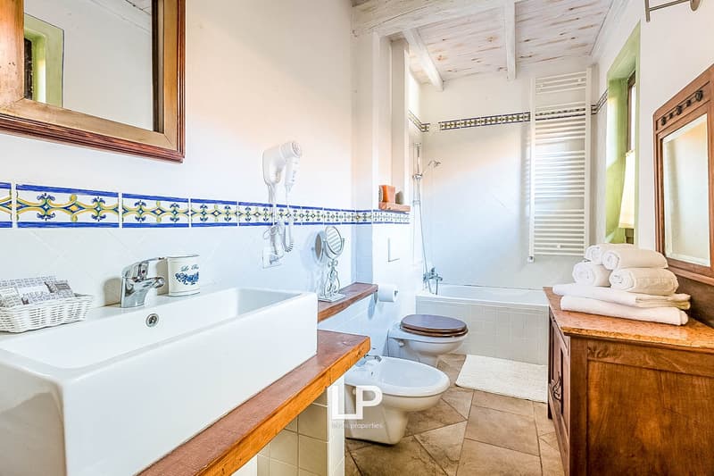 Spacious bathroom with white tile floor, wooden vanity with sink, toilet, and bathtub. Decorative tile border along the walls. Towels and other bathroom accessories visible.