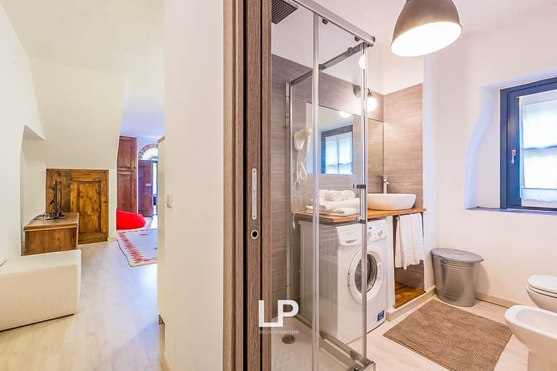 Modern bathroom with glass shower enclosure, vanity with sink, washing machine, and toilet. Wooden accents and neutral color scheme.