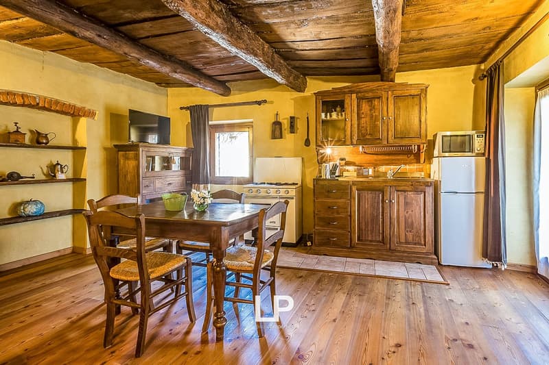 This image shows a rustic and cozy kitchen and dining area. The room features exposed wooden beams on the ceiling, giving it a charming, traditional feel. The walls are painted a warm yellow color, and the floors are made of wood planks. The kitchen area