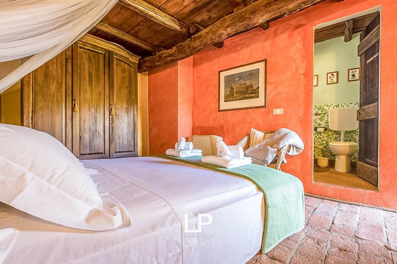 Rustic bedroom with wooden beams, orange walls, large wooden wardrobe, white bedding, and a fireplace visible in the bathroom area