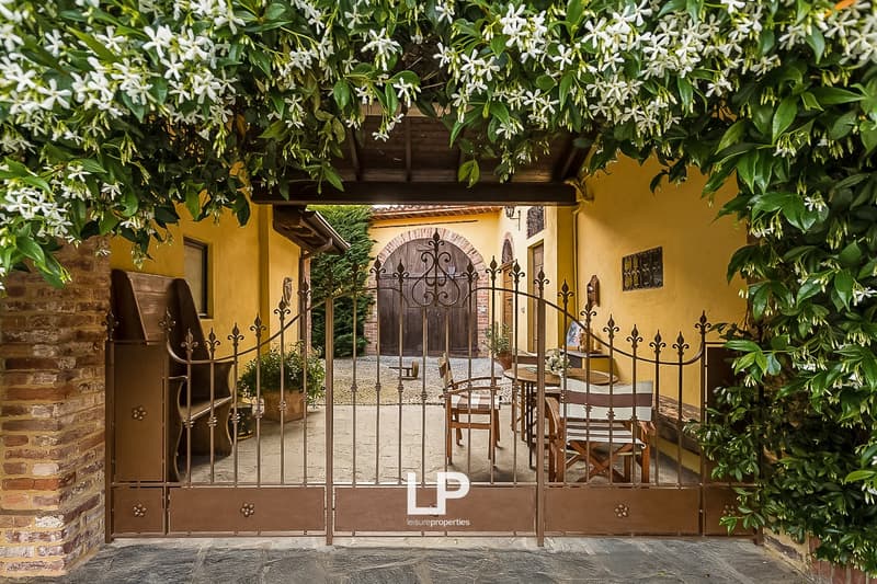 Charming stone house with a wrought iron gate, surrounded by lush greenery and flowering plants. The entrance features a decorative archway and a covered patio area.