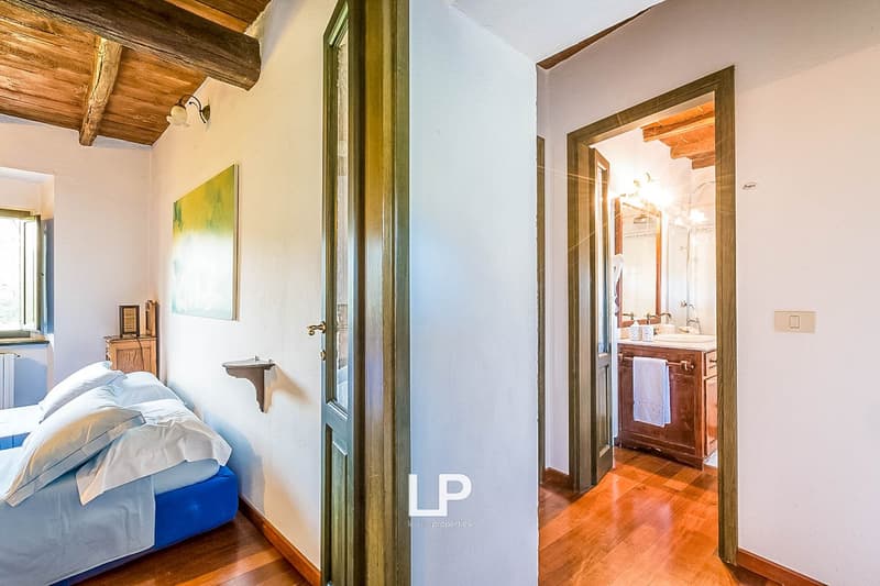 Bedroom with wooden ceiling beams, white walls, large window, bed, and private bathroom visible through the doorway