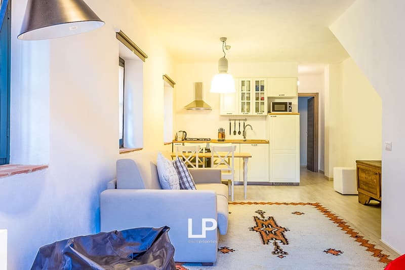 Open-concept kitchen and living room with white walls, hardwood floors, and modern appliances. The kitchen features a range hood, cabinets, and a dining table. The living room has a sofa and a patterned rug. There is also a microwave and a refrigerator vi
