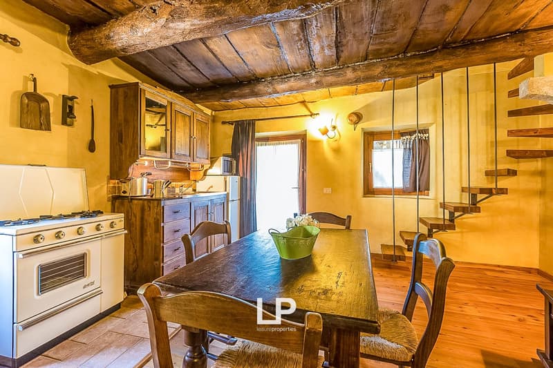 Rustic kitchen and dining area with exposed wooden beams, wooden cabinets and furniture, gas stove, and a wooden dining table with chairs. The room has a warm, cozy atmosphere with yellow lighting.