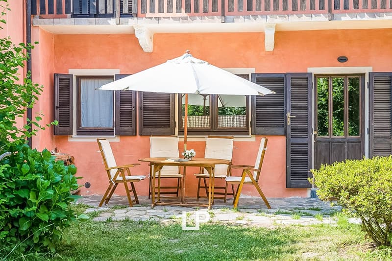 Outdoor patio with a wooden table and chairs, a white umbrella, and a grassy area in front of a pink stucco building with black shutters.