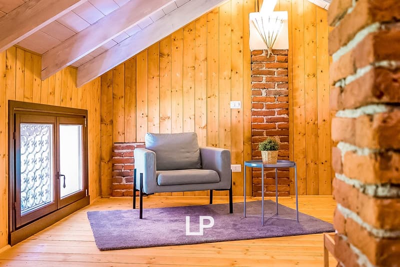 Cozy living room with wooden walls and ceiling, exposed brick fireplace, gray armchair, side table, and potted plant