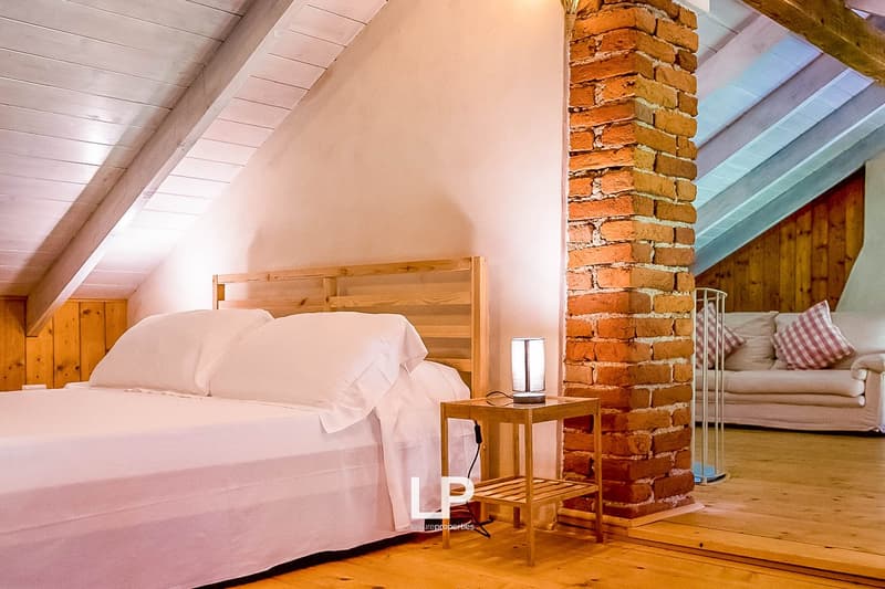 Cozy bedroom with wooden walls and ceiling, a brick fireplace, a wooden bed frame with white bedding, and a small side table.