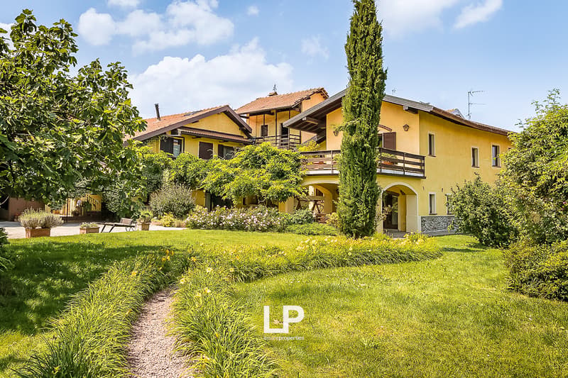 2-story yellow house with red tile roof, surrounded by lush greenery including trees, shrubs, and a well-manicured lawn. The house has balconies and a parking area on the property.