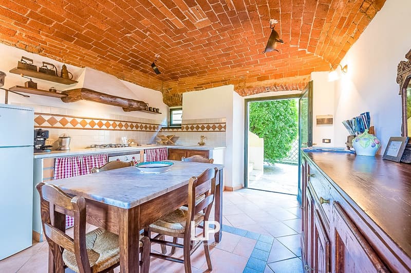 Rustic kitchen with brick vaulted ceiling, wooden dining table and chairs, tiled floor, kitchen appliances, and a view of the outdoor garden through the open doorway.