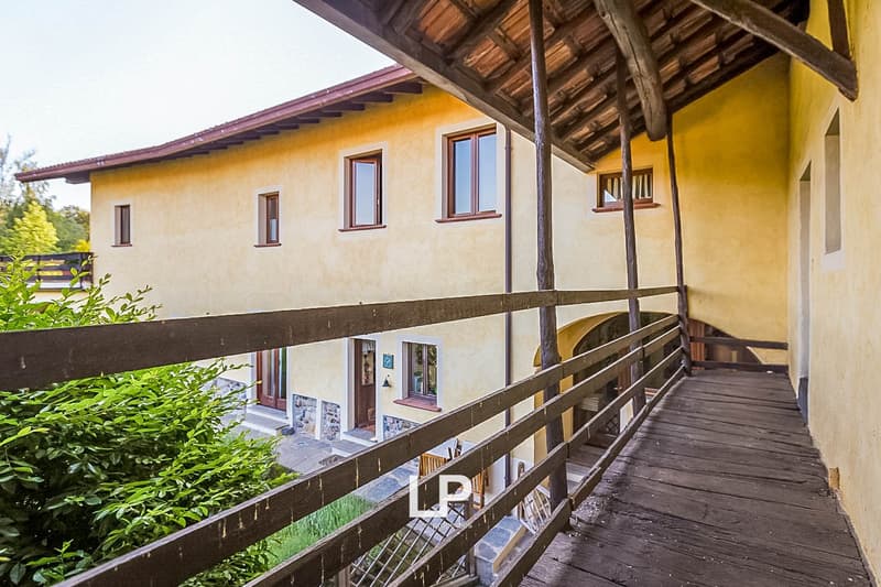 2-story house with yellow exterior walls, wooden roof, and a wooden balcony/terrace with a railing. The house has several windows and is surrounded by greenery.